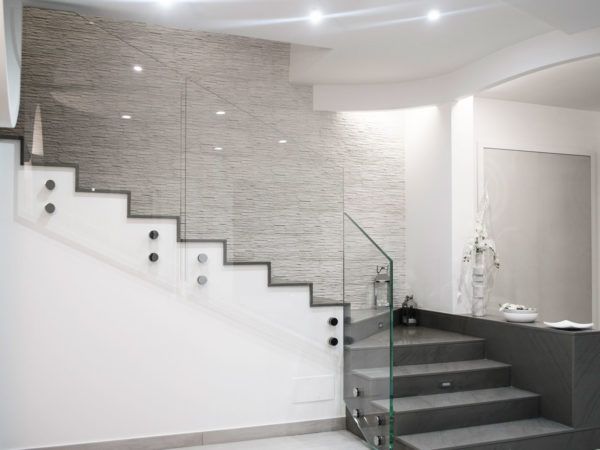Scala moderna con ringhiere in vetro, gradini grigi e pareti bianche. Dotata di illuminazione da incasso e parete decorativa in pietra testurizzata.