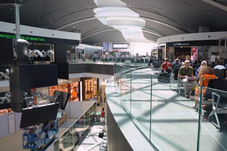 Interno di un terminal aeroportuale con negozi e un'area salotto, ringhiere in vetro e luce naturale proveniente da un grande lucernario.