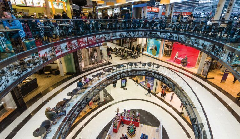 Interno di un centro commerciale a più livelli con balconi curvi, persone che camminano e curiosano e una forte illuminazione dall'alto.