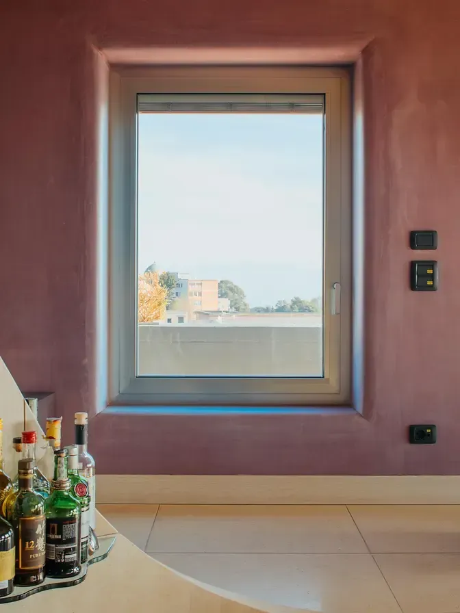 Finestra con vista panoramica. Pareti rosa, cornice della finestra marrone chiaro e un bancone con bottiglie di liquore in primo piano.