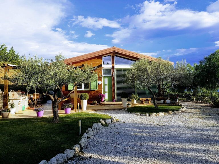 Una casa in legno con un vialetto di ghiaia, ulivi e persiane verdi, che si staglia contro un cielo azzurro.