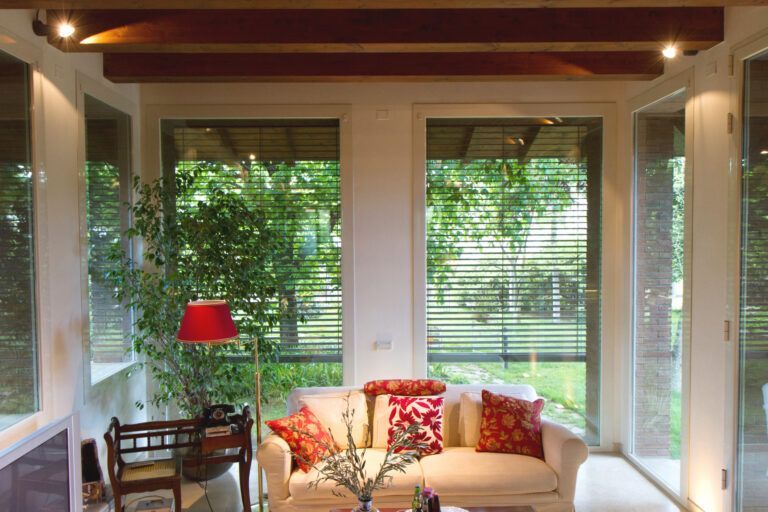 Accogliente veranda con divano bianco, cuscini rossi e ampie finestre con vista sul giardino verde. Travi in legno e illuminazione a incasso.