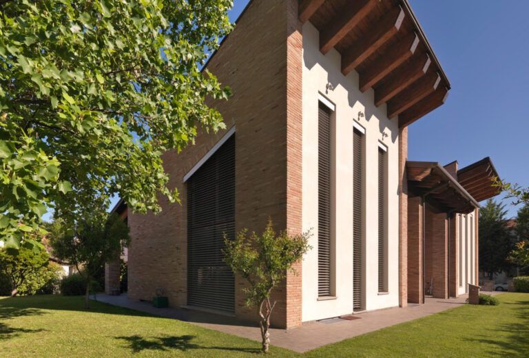 Moderna chiesa in mattoni e stucco, con alte finestre e tetto spiovente, su un prato erboso con alberi, sotto un cielo azzurro.