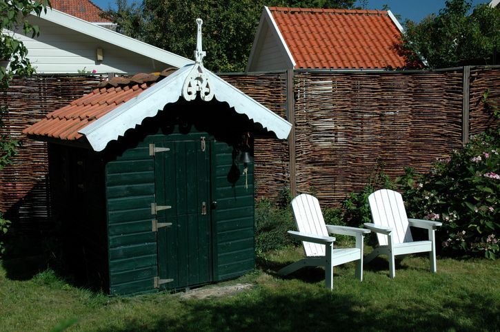 Capanno verde con finiture bianche e tetto di tegole rosse, due sedie Adirondack bianche in un giardino soleggiato.