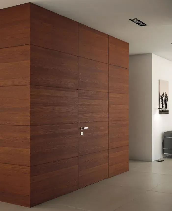 Una parete rivestita in legno con una porta nascosta. Pannelli in legno marrone formano una porta a filo con una maniglia argentata. L'ambiente è moderno e minimalista.