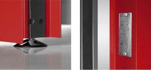 Primo piano di una porta rossa con finiture nere. Una piccola piastra metallica è rivettata alla porta e un piede nero è appoggiato sul pavimento.