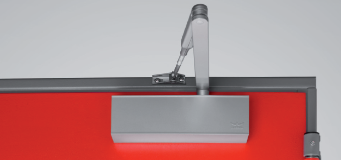 Primo piano di un chiudiporta montato su una porta rossa, con telaio grigio e parete bianca.