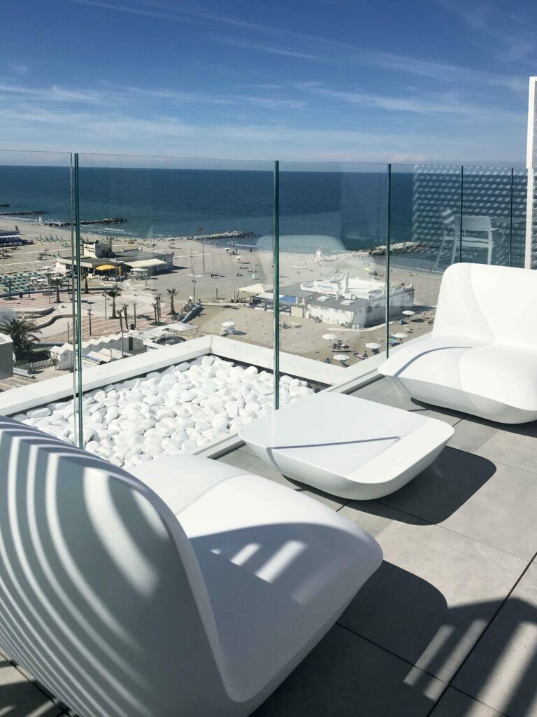 Patio sul tetto con mobili bianchi, pietra bianca e ringhiera in vetro trasparente con vista sulla spiaggia e sull'oceano in una giornata di sole.