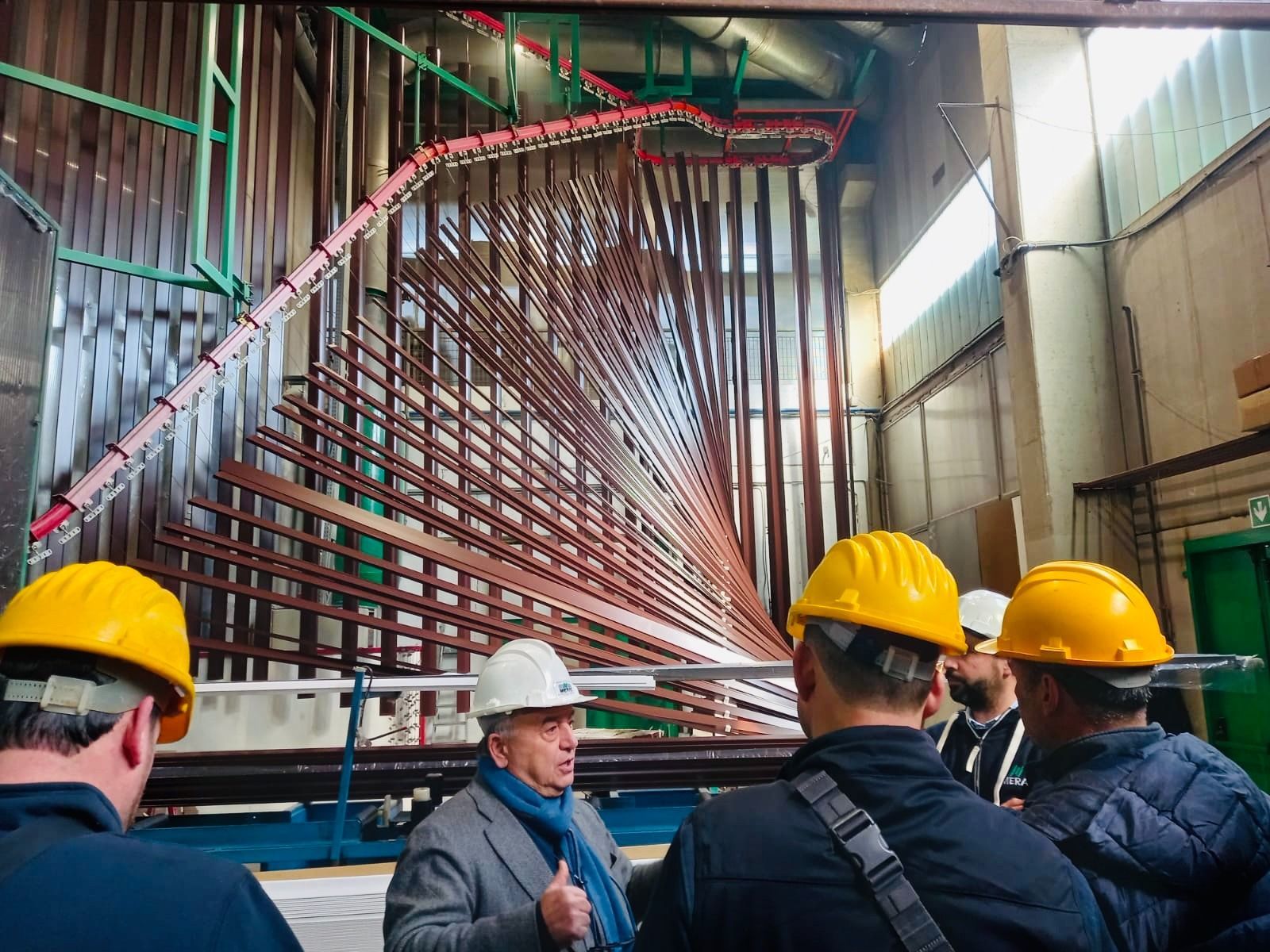 Un gruppo di persone con il casco osserva un grande macchinario industriale con numerosi tubi di rame. Si trovano all'interno di una fabbrica.