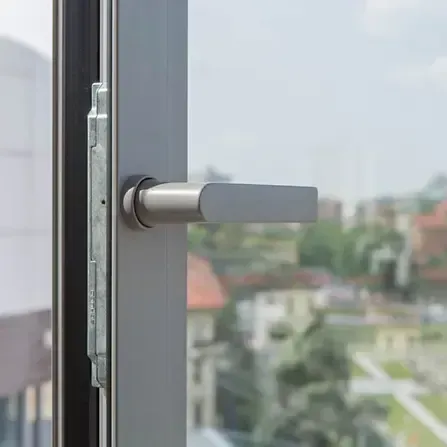 Primo piano della maniglia di una finestra, metallo argentato su una cornice grigia, con un paesaggio urbano sfocato sullo sfondo.