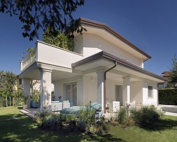 Casa bianca a due piani con veranda e balcone, immersa in un prato verde sotto un cielo azzurro.