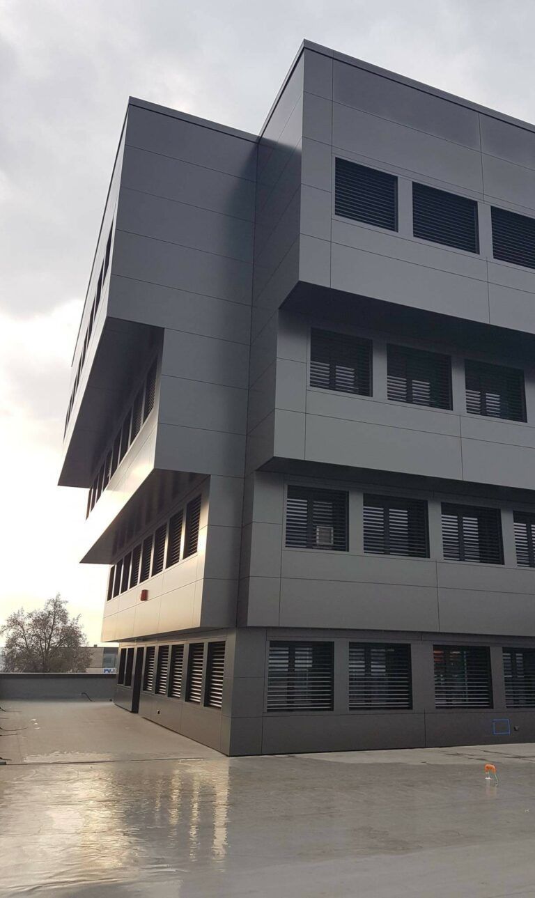 Edificio moderno grigio con finestre rettangolari e piani superiori sporgenti. Cielo nuvoloso sopra.