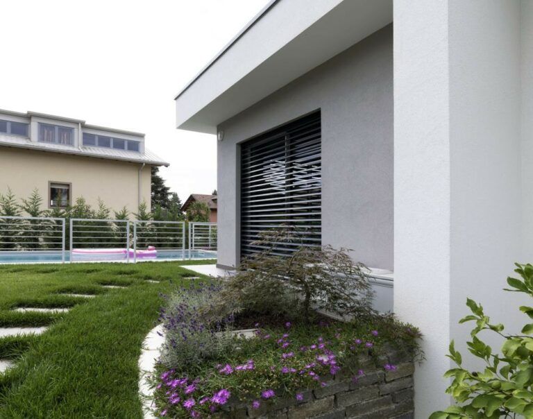 Esterno di una casa moderna con un'aiuola in pietra e un prato verde. Sono visibili una finestra con persiane oscuranti e una piscina.