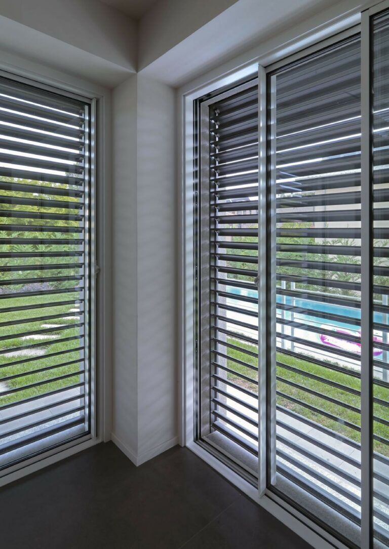 Finestre angolari con persiane grigie, che si affacciano su un'area erbosa e su una piscina esterna.