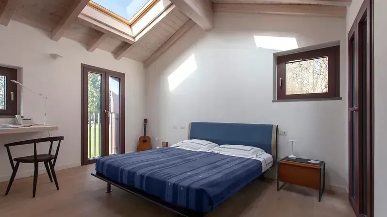 Camera da letto luminosa con letto blu, scrivania e lucernario sul soffitto, vicino alla porta che conduce a un balcone.