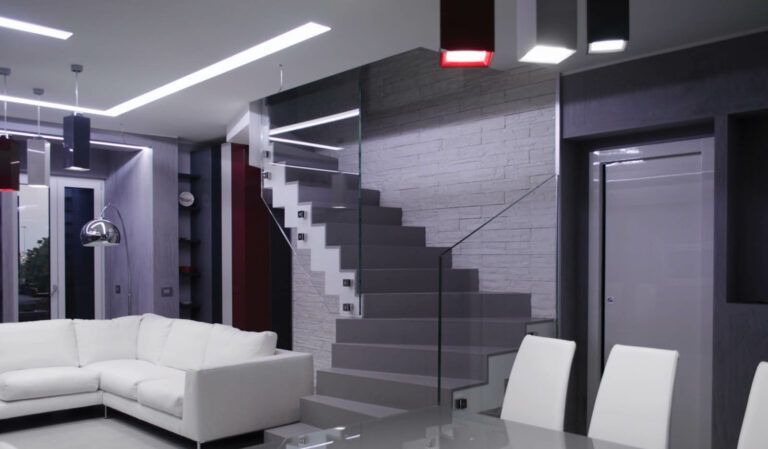 Interni moderni con divano componibile bianco, scala grigia e ringhiera in vetro. Tavolo da pranzo e sedie in primo piano, con luci sospese.