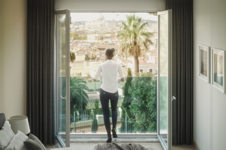 Donna in piedi davanti alle porte-finestre aperte, che guarda un paesaggio urbano con palme. Indossa una camicia bianca e pantaloni neri.