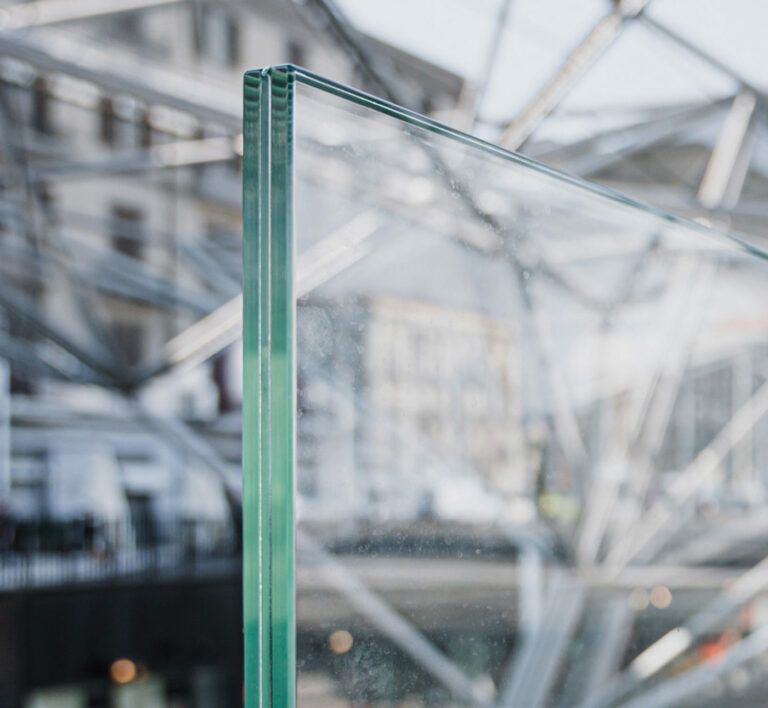 Primo piano del bordo di due pannelli di vetro trasparente, probabilmente una finestra isolata, sullo sfondo della facciata sfocata di un edificio moderno.