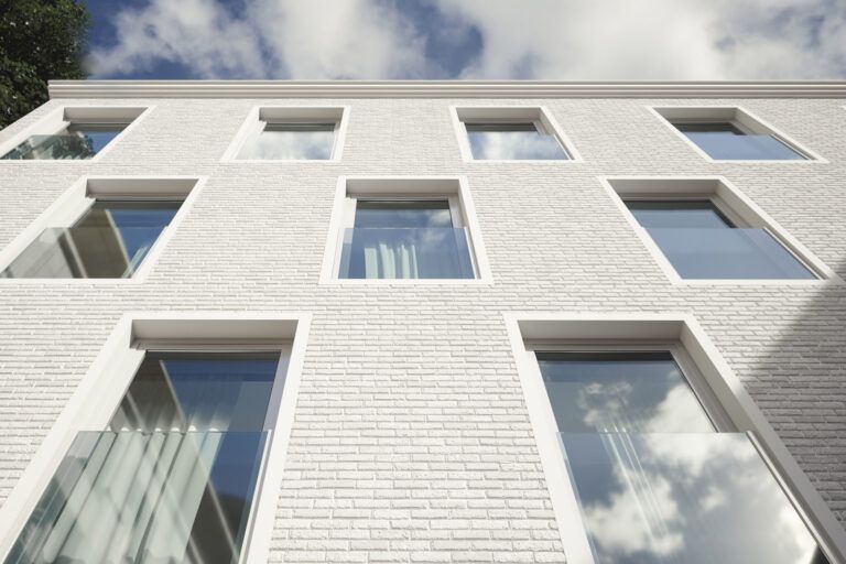 Edificio in mattoni bianchi con nove finestre che riflettono il cielo azzurro. Alcune finestre hanno balconi in vetro.