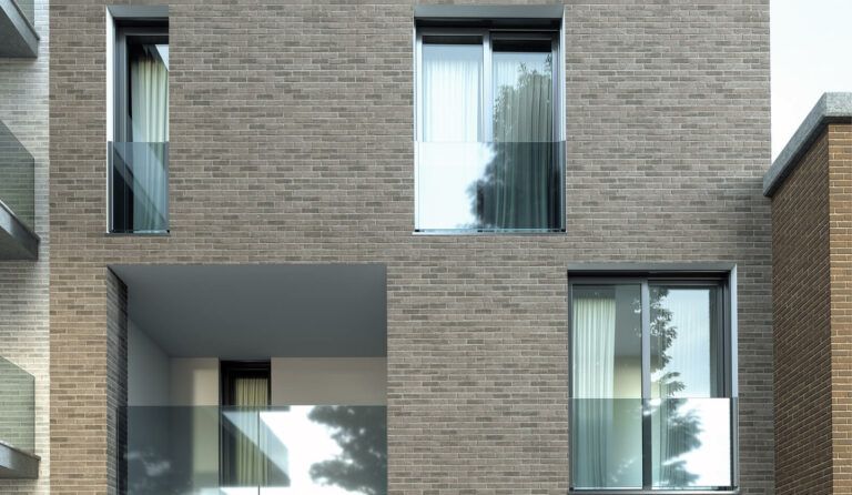 Un moderno edificio in mattoni con tre finestre e un balcone all'aperto. Le finestre hanno persiane bianche e sul vetro si vede il riflesso di un albero.