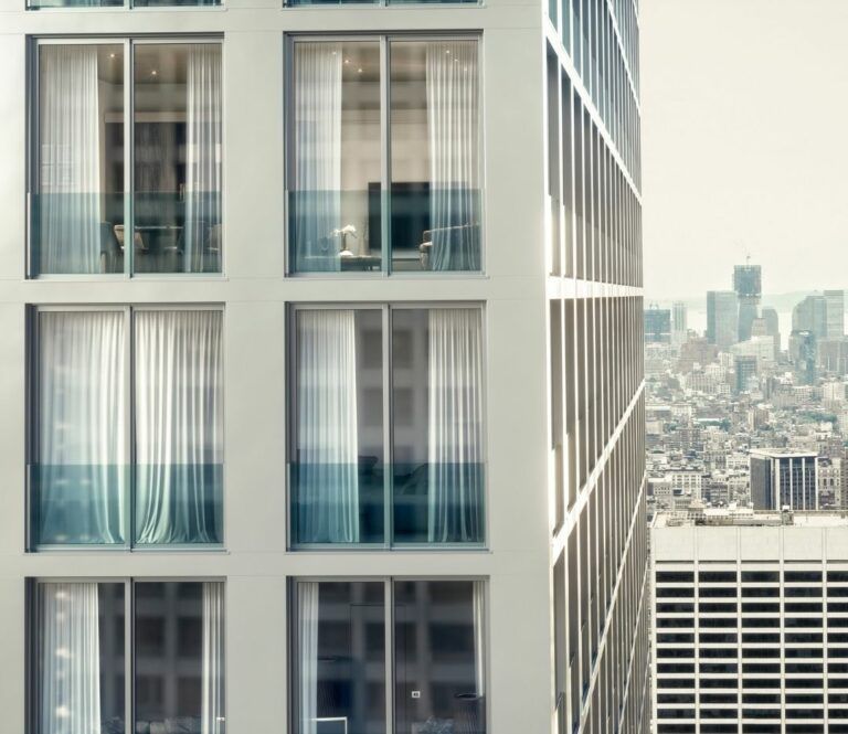 Esterno di un edificio alto con ampie finestre e tende bianche che si affacciano sul paesaggio urbano.