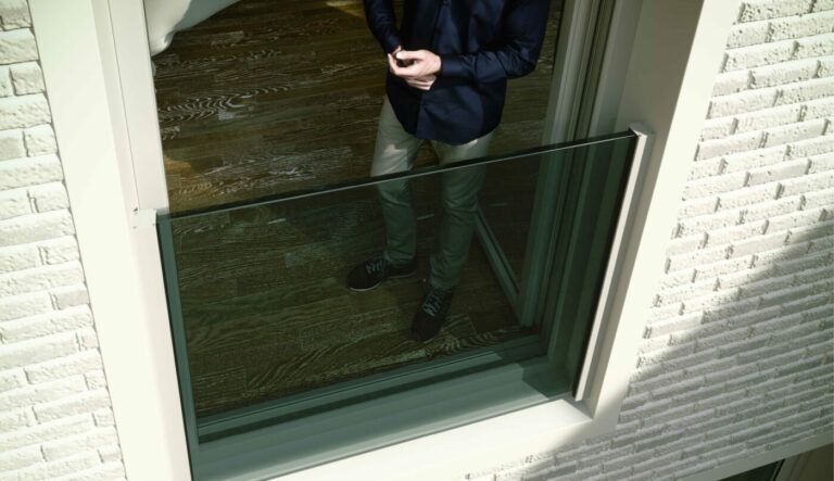 Persona in piedi all'interno di un edificio, visibile attraverso una grande finestra aperta. Indossa una camicia blu e pantaloni grigi. Esterno in mattoni bianchi.