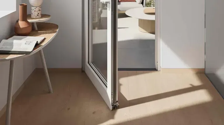 Un interno con pavimento in legno chiaro e una porta a vetri aperta che conduce a uno spazio esterno soleggiato, arredato con un tavolino, un libro e un vaso.