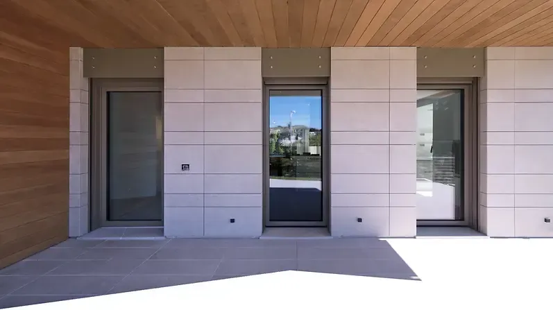 Vista esterna di un edificio con tre porte in vetro incorniciate da pannelli in pietra chiara sotto un soffitto in legno.