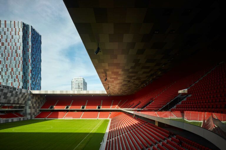 Una vista dall'interno di un grande stadio rosso con gradinate e un campo verde, sotto una copertura parziale, con un edificio moderno sullo sfondo.