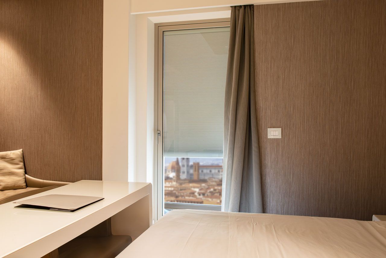 Camera da letto con scrivania bianca, letto e finestra con persiane e tenda grigia. Le pareti sono di un marrone testurizzato e la vista esterna include edifici.