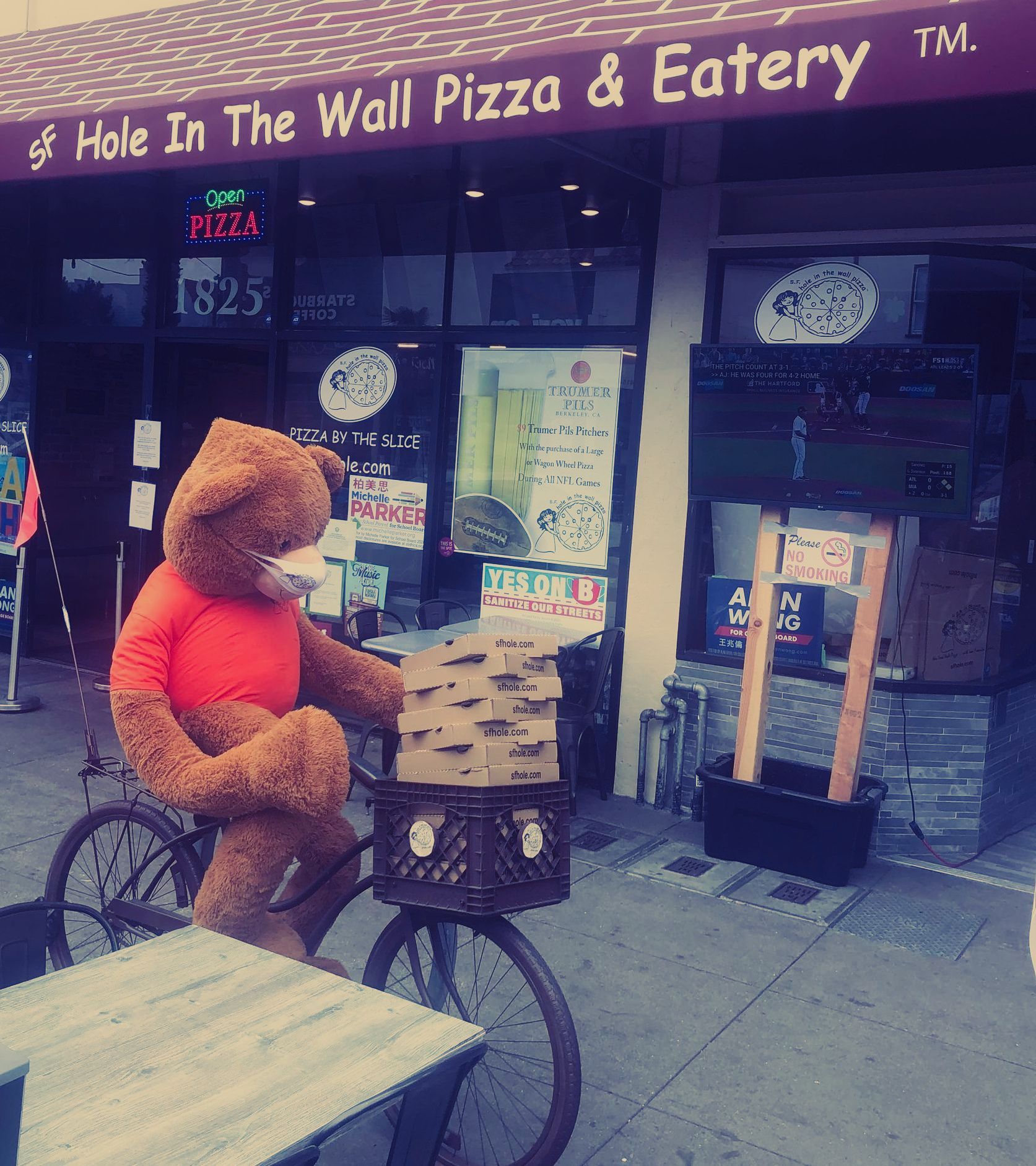A teddy bear is riding a bike in front of a hole in the wall pizza and eatery