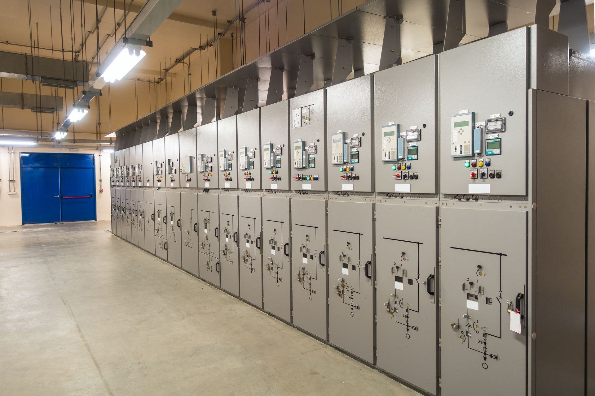 Row of gray electrical panels in an industrial setting, with overhead lighting and a blue door.