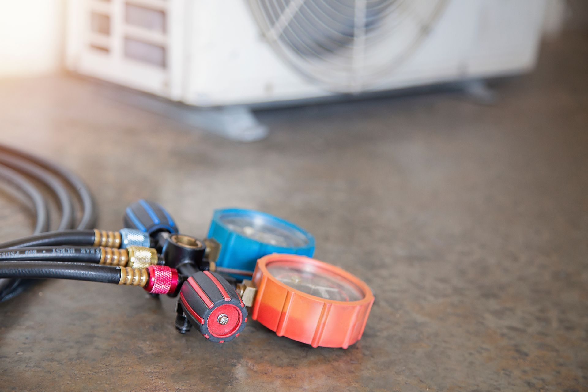 Close-up of HVAC manifold gauge set with hoses on a concrete floor near an outdoor unit.