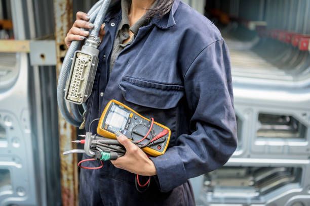 A person in a blue jumpsuit holding a multimeter and cables.