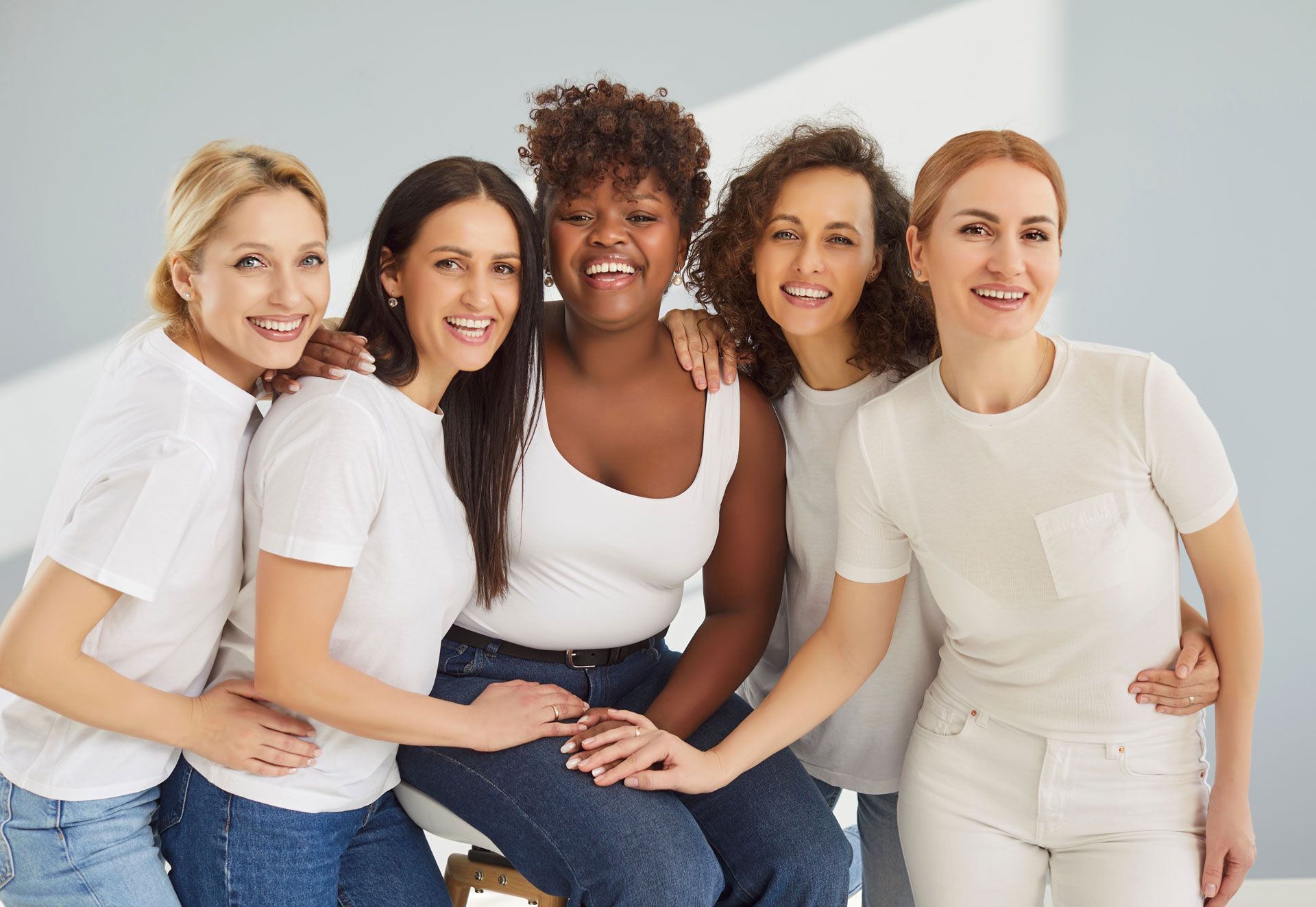Five smiling people in white tops stand together with their hands joined in a supportive gesture against a gray background.