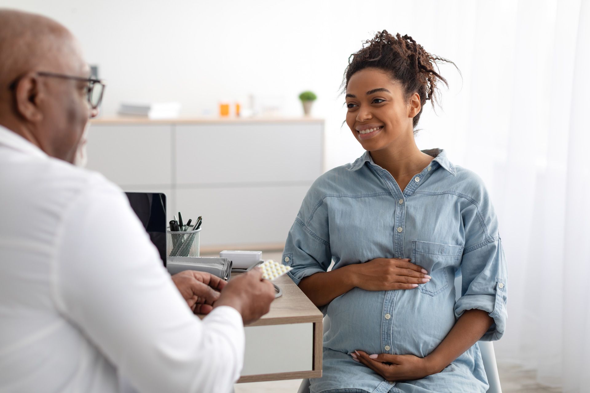 Pregnant woman smiles at a doctor holding medicine in a light-filled medical office.