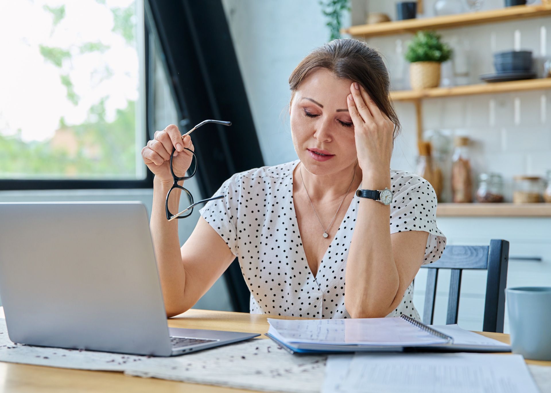 Woman with headache, holding glasses, working at laptop in kitchen.