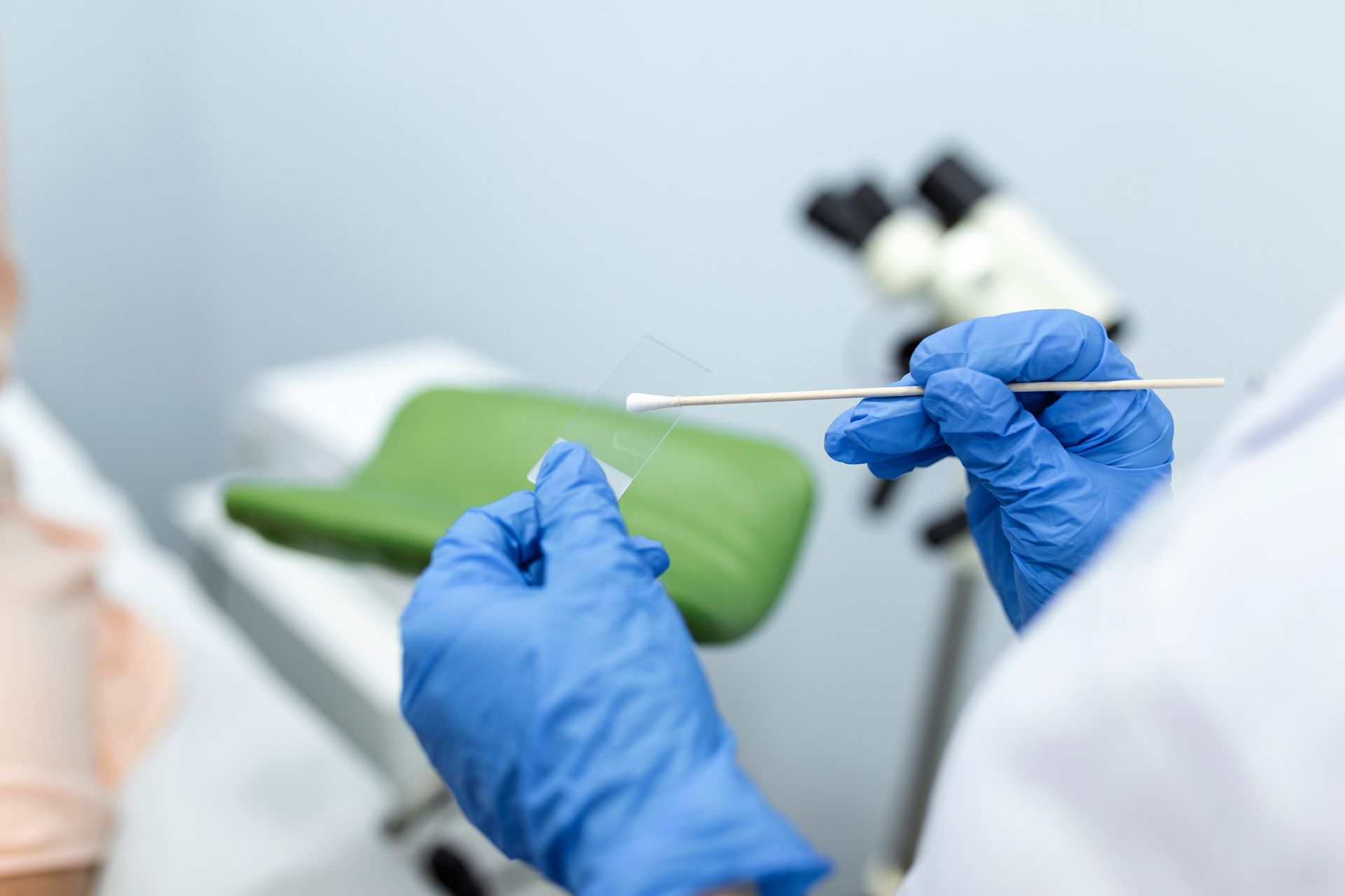 Medical professional in blue gloves holding a swab and sample slide near a microscope.