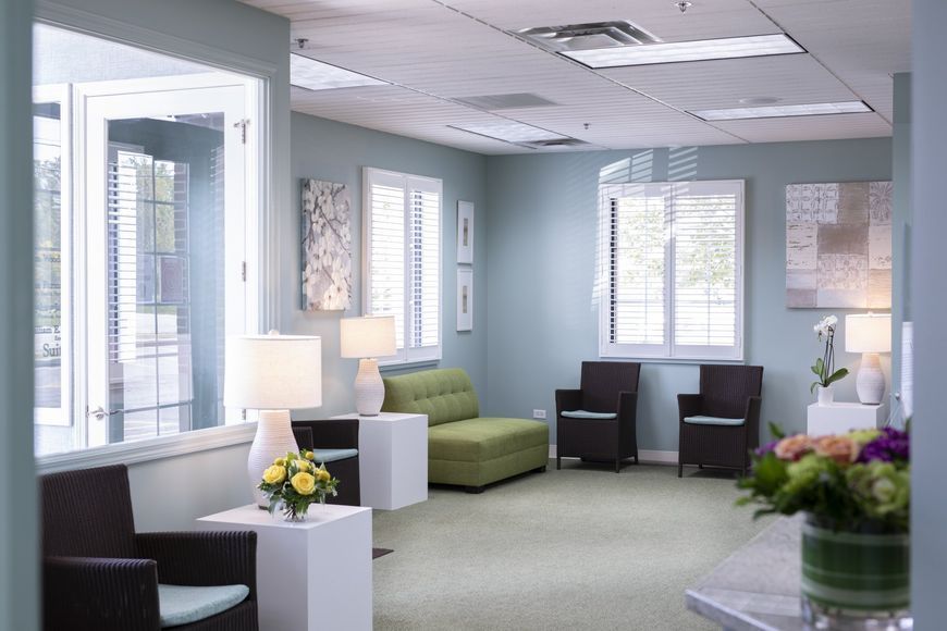 A waiting room with blue walls, windows, chairs, and a green couch, flowers.