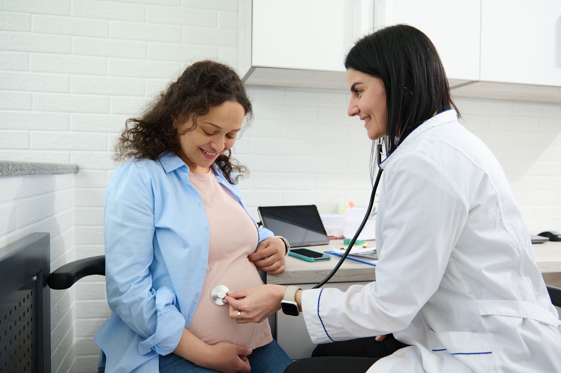 Pregnant person being examined by a doctor with a stethoscope in a medical office.