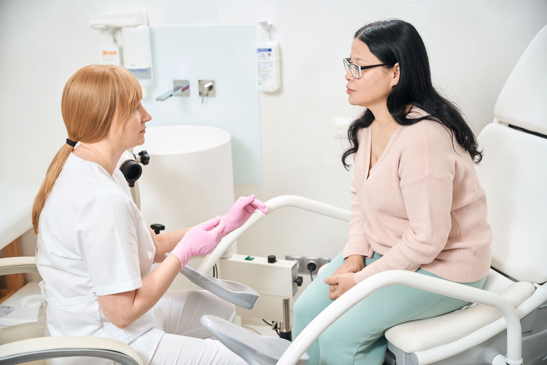 Doctor speaking to a patient in an examination room. The patient sits in a chair, while the doctor wears gloves.