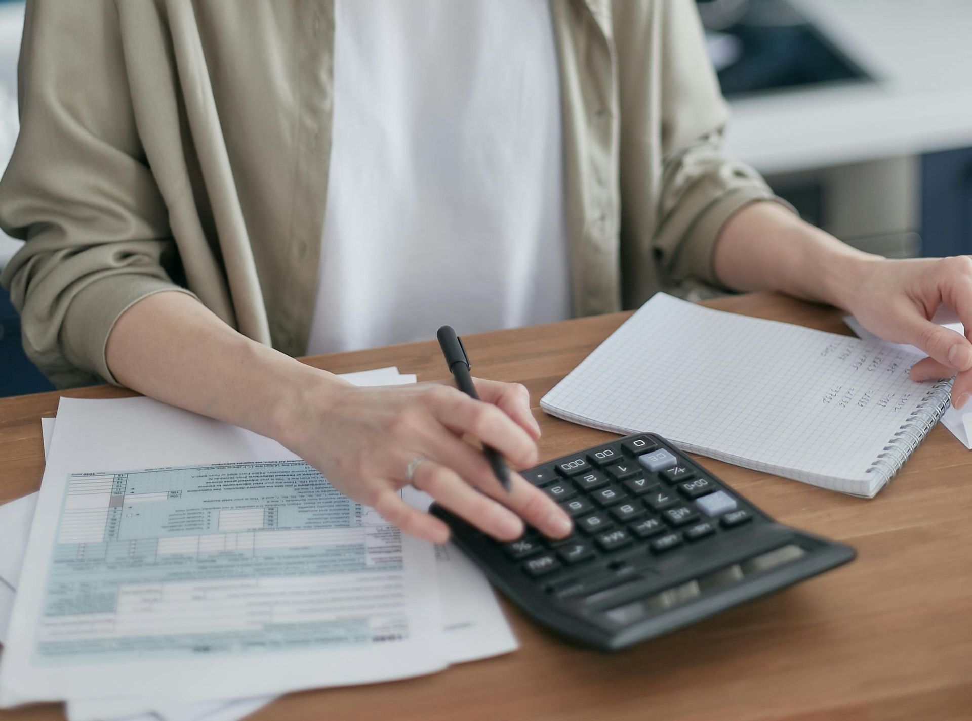 A woman is sitting at a table using a calculator and writing in a notebook.