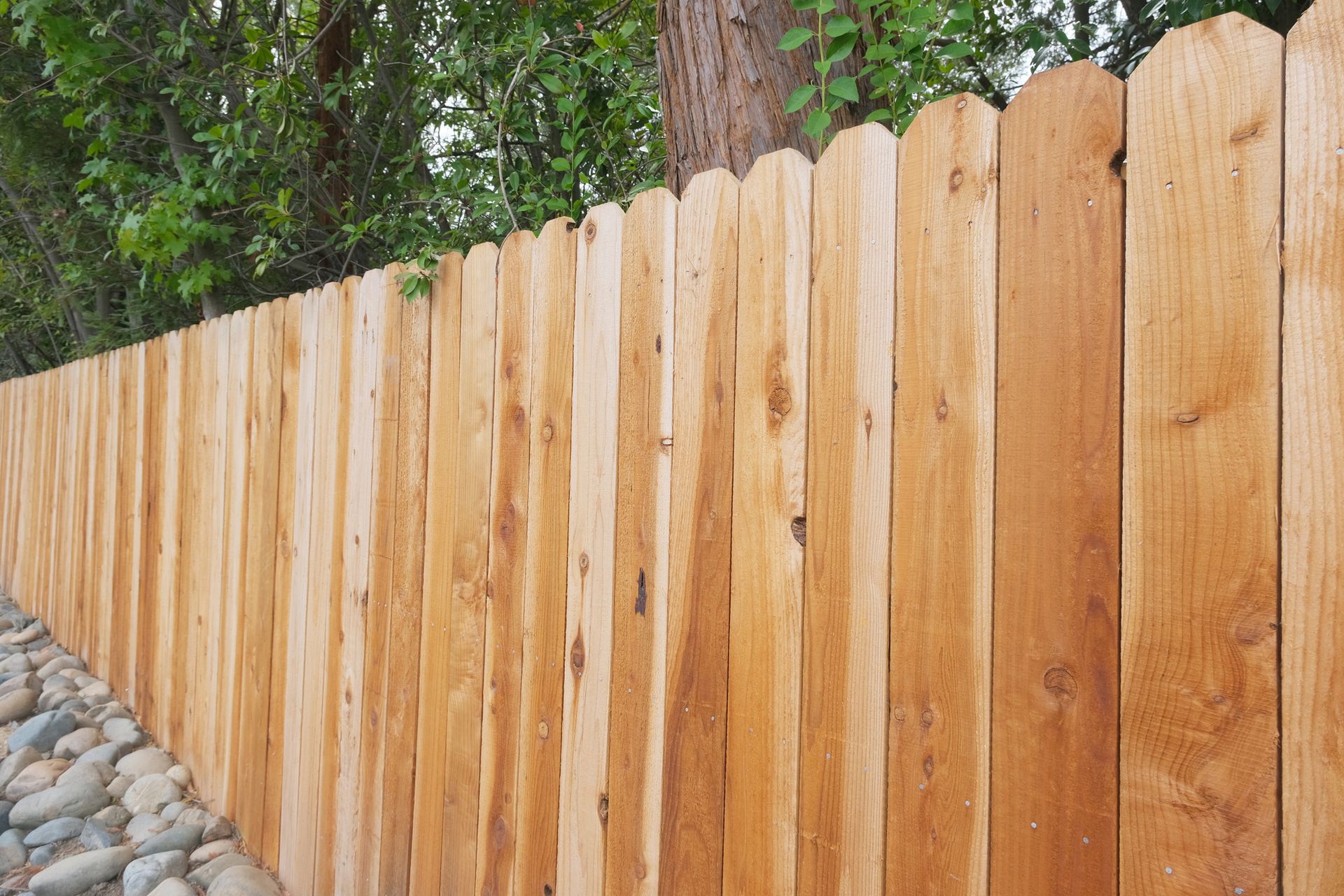 Wooden fence with scalloped top, built next to a rocky ground, with trees in the background.