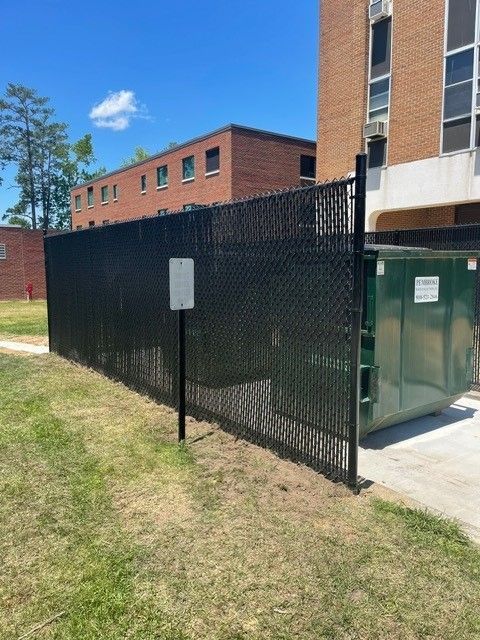 A black chain link fence surrounds a dumpster in front of a brick building.