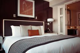 A hotel bedroom features a large bed with several pillows and a patterned throw. A painting and a doorway to a bathroom are visible.