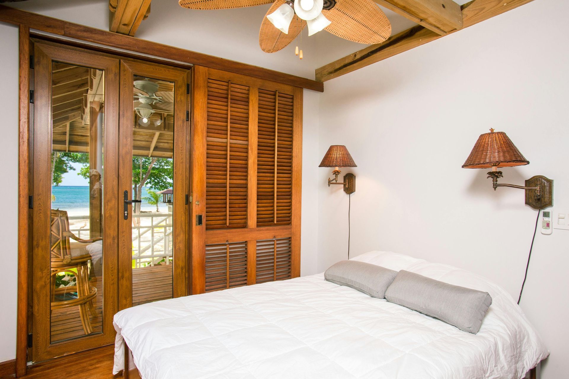 Bedroom with wooden doors leading to a beachfront view, bed with gray pillows, and woven lamps.