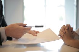 Man in suit presenting documents to a person with hands clasped, likely a business meeting or signing of a contract.
