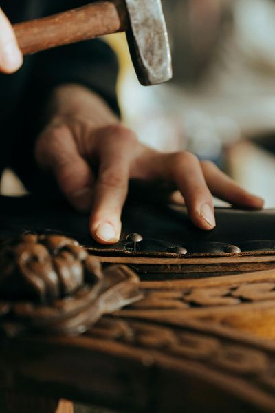 A person hammering a small nail into a wooden surface, likely for woodworking. Close-up of hands and tool.