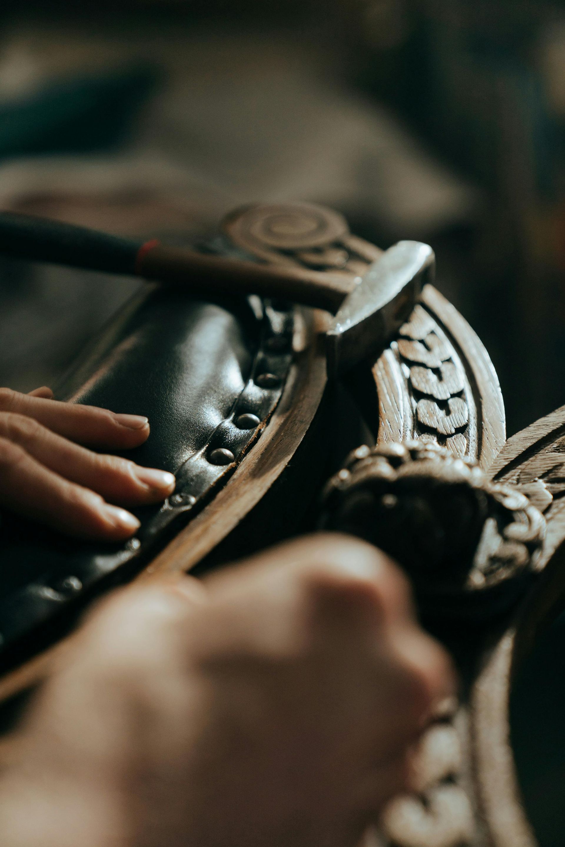 A craftsman's hands working on a dark leather piece, hammering rivets into place on a carved wooden chair.
