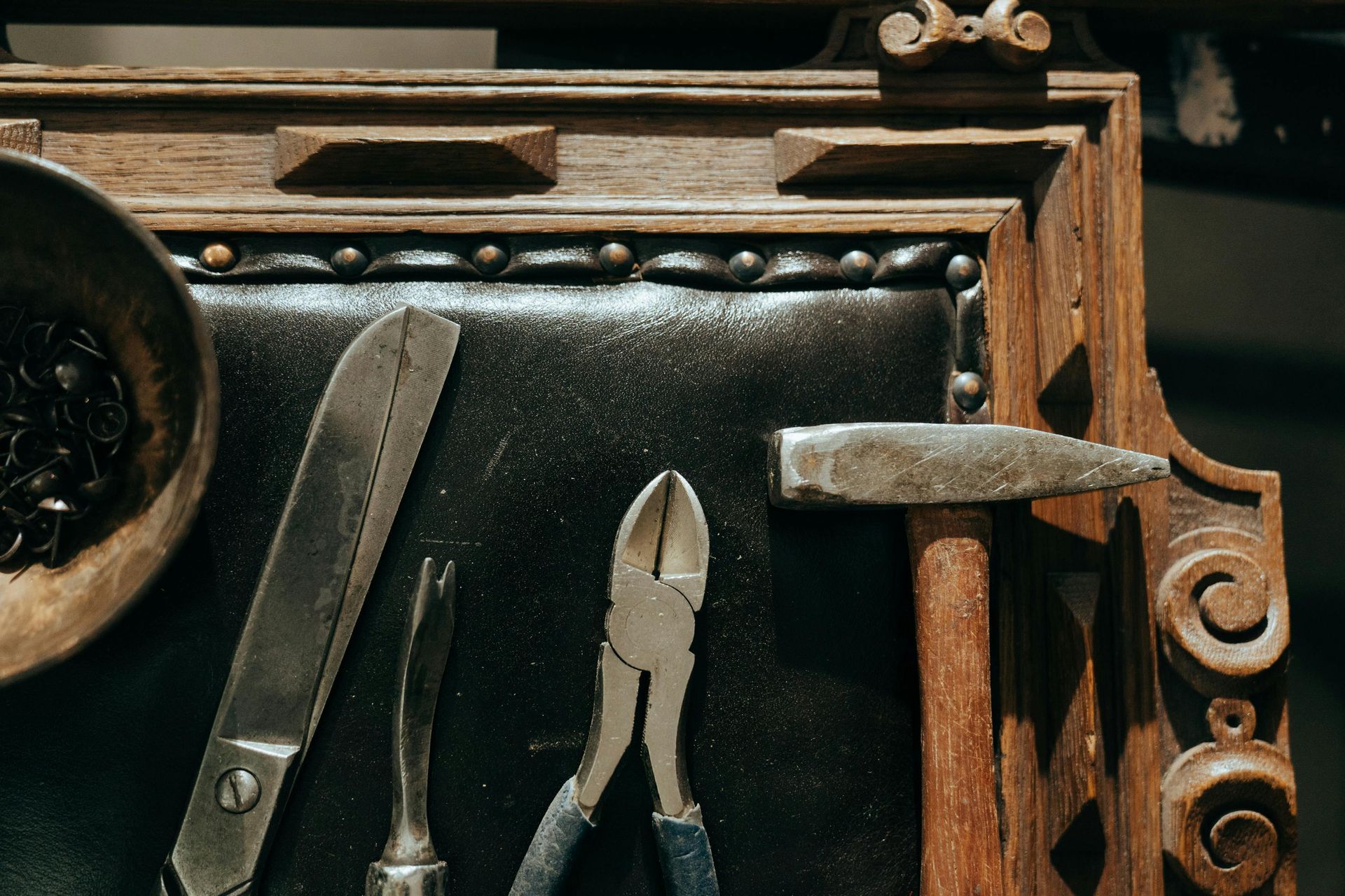 Tools on a wooden framed workspace: scissors, a curved tool, pliers, a hammer, and a bowl of metal rings.